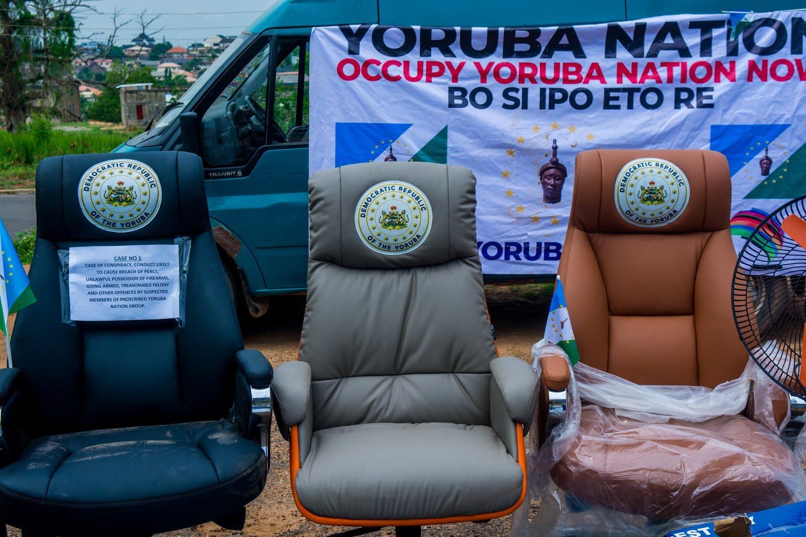 Three empty chairs in front of a van with a "Yoruba Nation" banner, symbolizing a protest or movement.