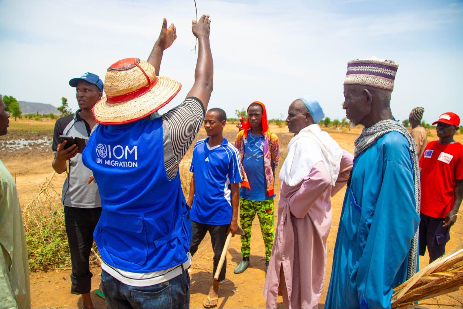 Group of people with a person in a blue UN Migration vest interacting with locals in a rural setting.