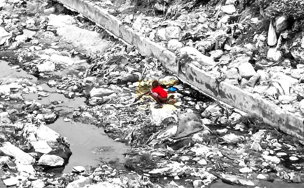 A selective color image of a person in red crouched amongst litter in a dry urban streambed.