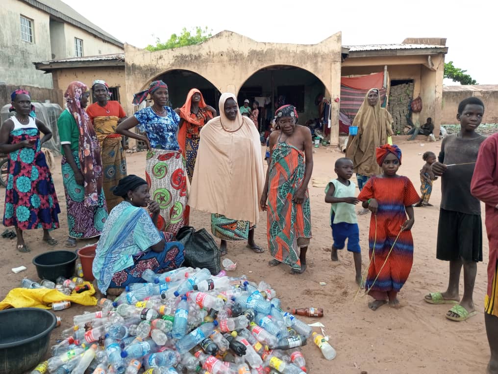 A group of people, including children, standing near a pile of plastic bottles in a village setting.