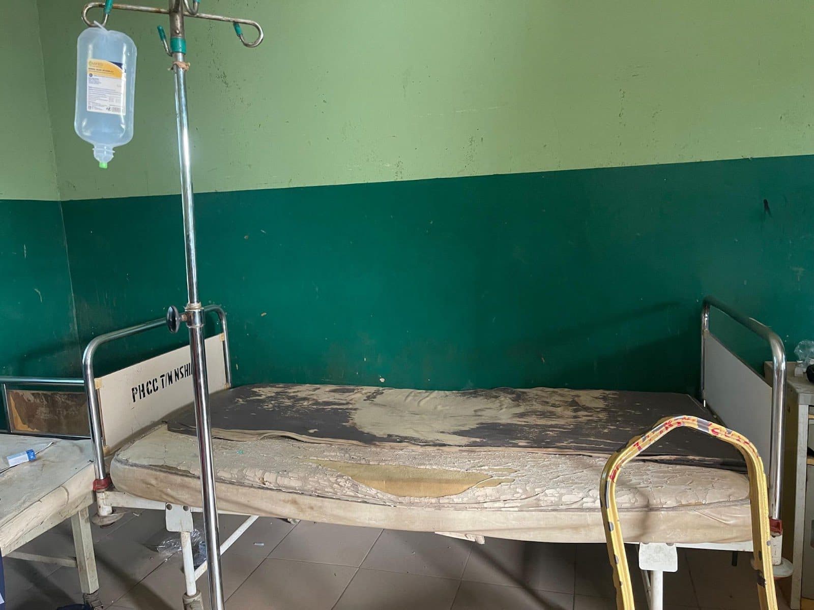 Worn hospital bed with damaged mattress in a green-walled room, next to an IV stand.