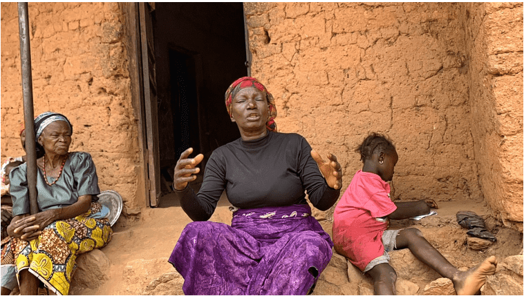 Three people sit outside a mud brick house. A woman gestures, another sits silently, and a child writes on paper.