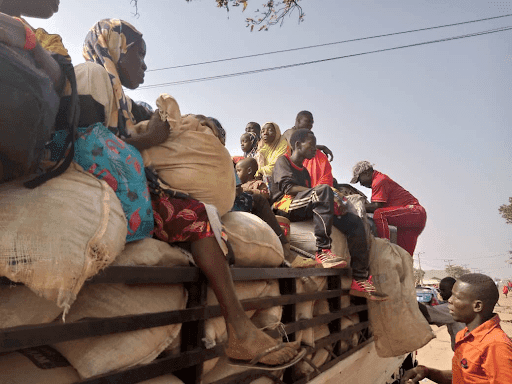People sitting on a loaded truck with bags, heading to an unknown location. Bright daytime with clear skies.