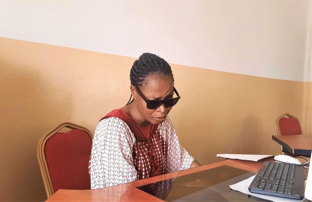 Scholastica Kalama sits at her desk at the National Centre for Women Development premises in Abuja. She is wearing dark shades and raising one eyebrow as she speaks. There is a desktop computer and keyboard in front of her. She is wearing a white and wine dress.