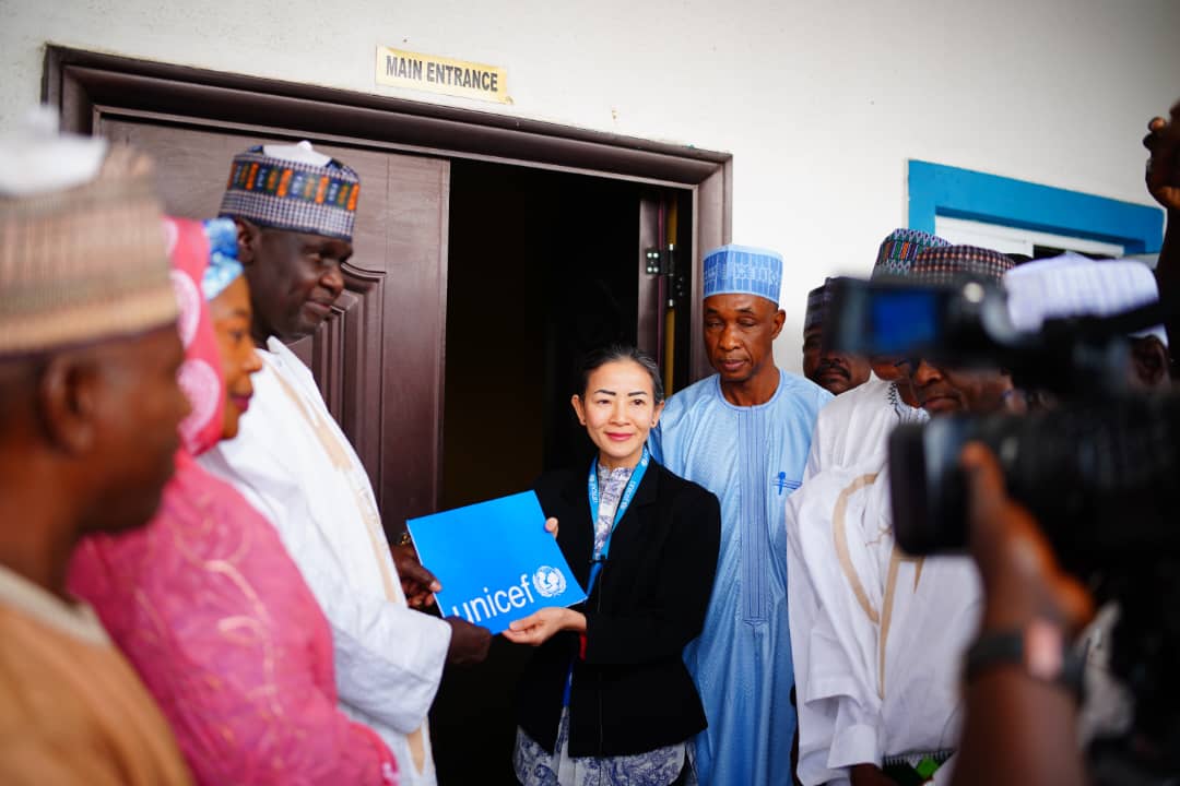 A group of dignitaries with a woman holding a UNICEF folder by a building's main entrance, with cameras pointing at them.