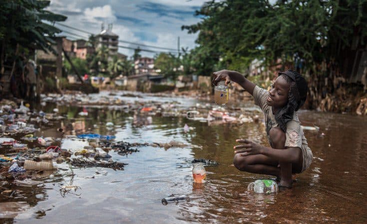 Girl squatting in polluted water, examining a bottle with murky liquid, surrounded by trash.