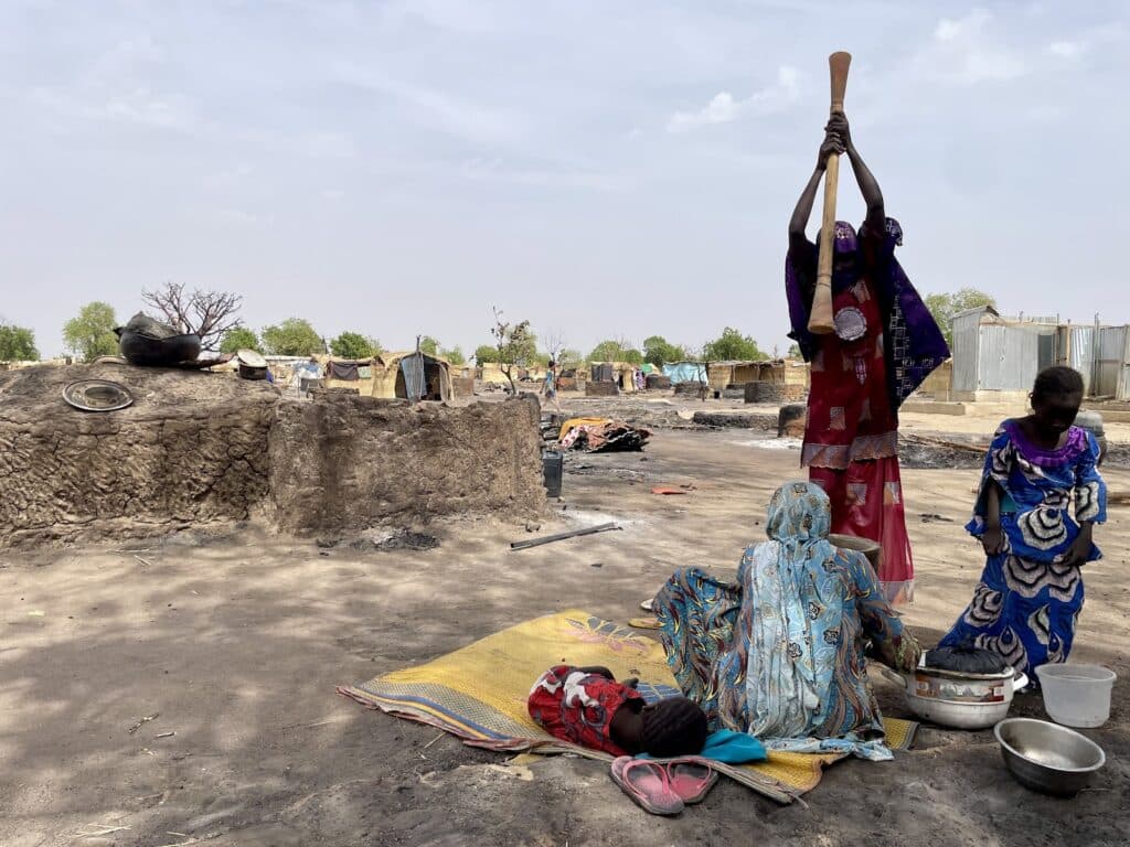Women and a child in traditional attire in a rural setting with huts and cooking utensils.