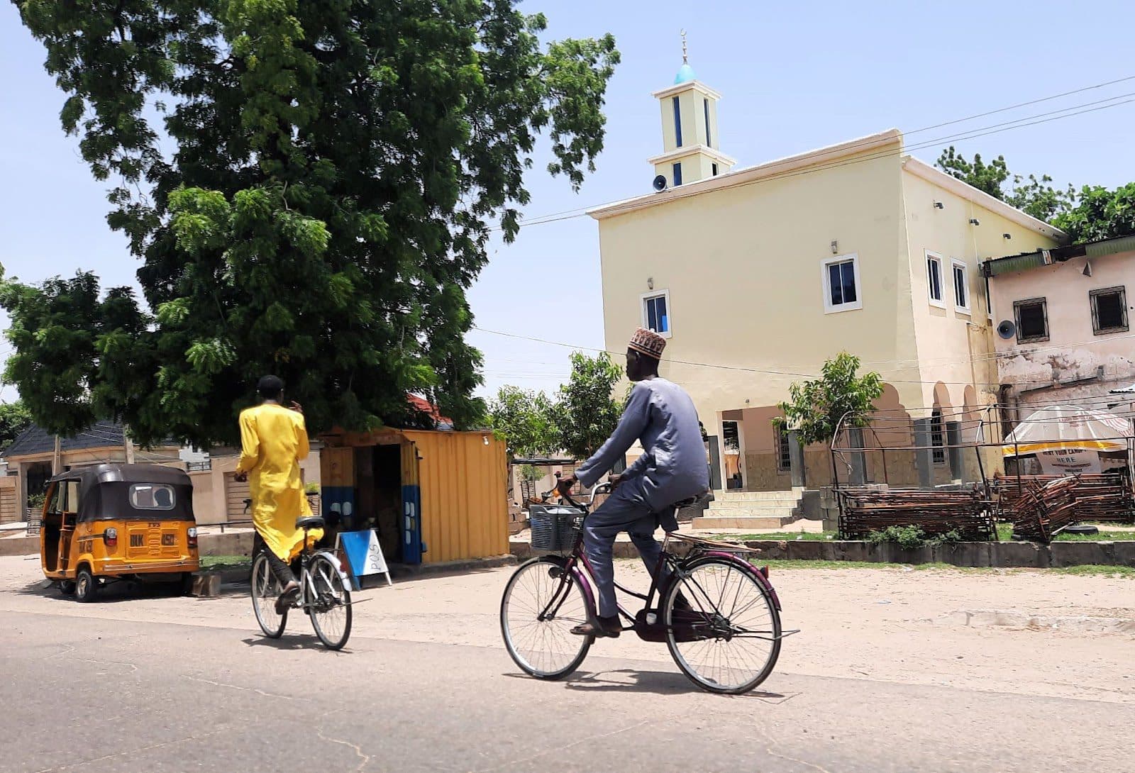 Two individuals on bicycles near a cream-colored building with a minaret, under a clear sky.