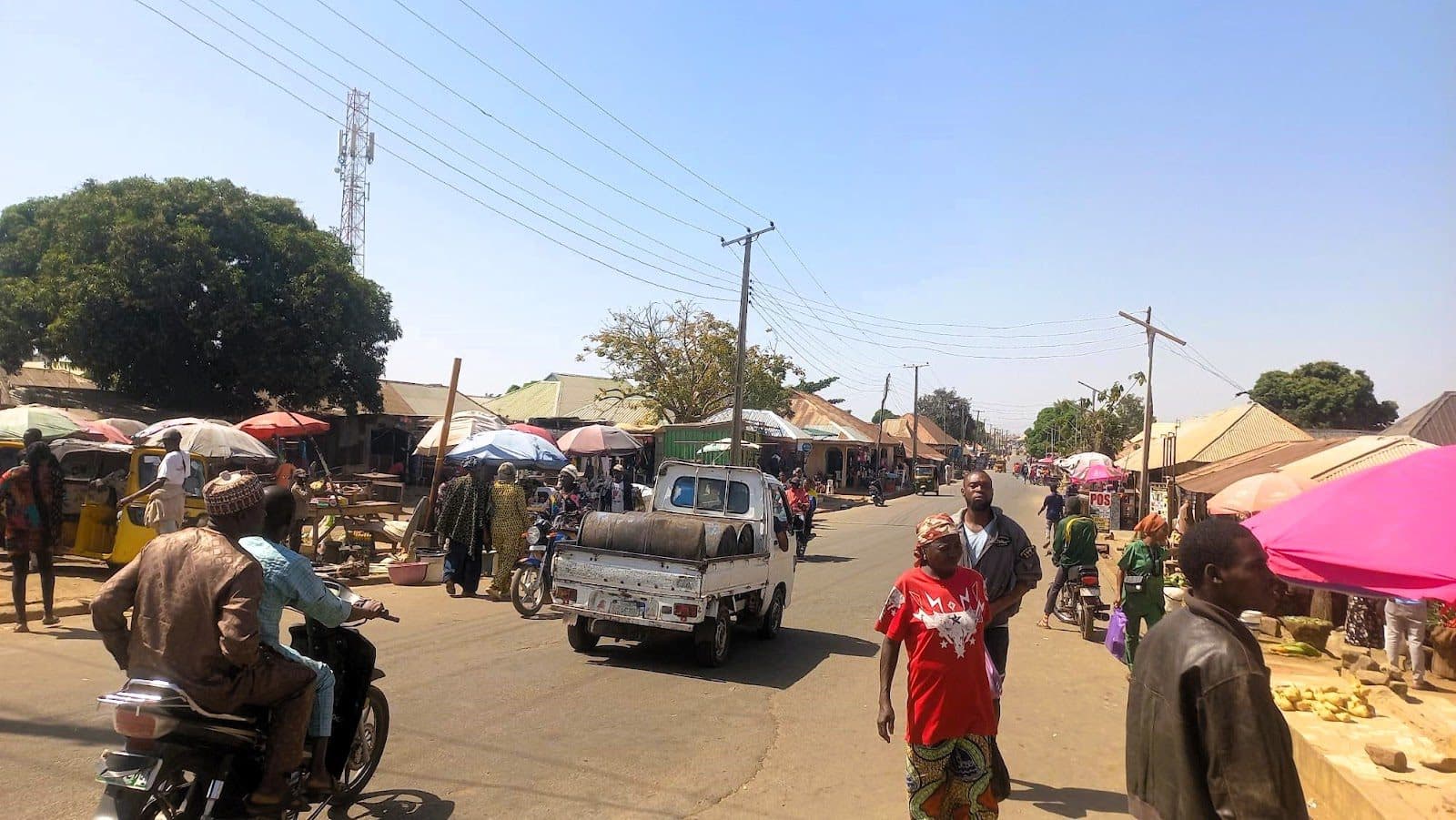 Busy street market scene with people, motorcycles, and a truck under a sunny sky.