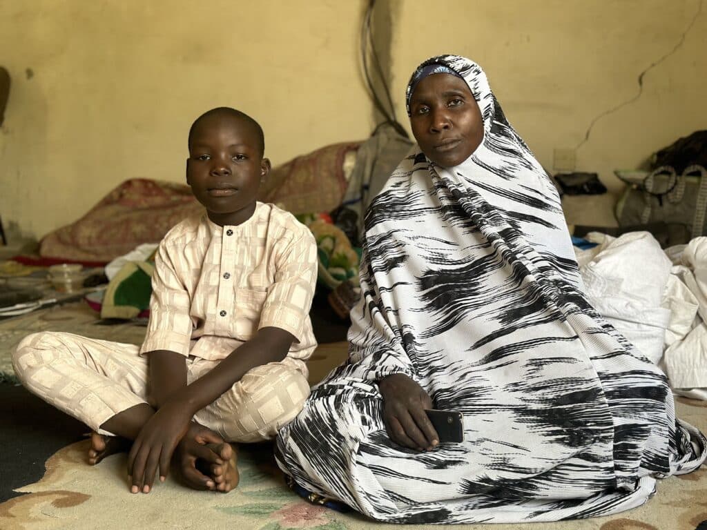 A boy and a woman sitting indoors with a mattress in the background, both looking calmly at the camera.