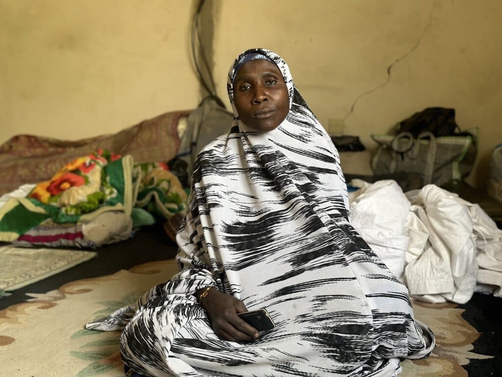 Woman in a black and white garment sitting in a modest room with textiles in the background.