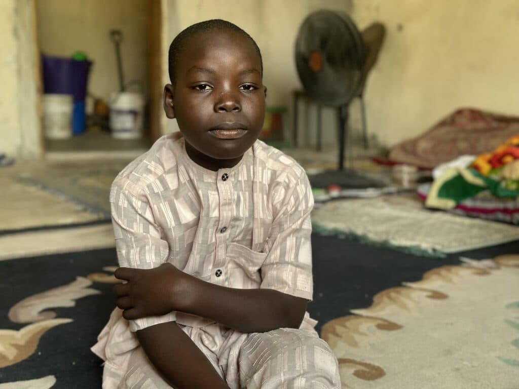 A young boy sits cross-armed in a room with a fan and colorful bedding in the background.