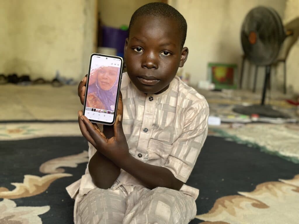 A young boy sitting indoors holds up a smartphone displaying a woman