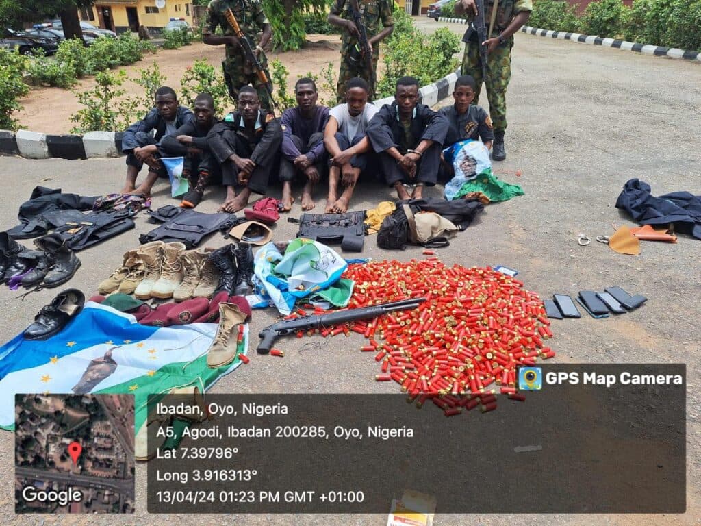 Group of men seated on the ground with soldiers standing, surrounded by clothes, shoes, and numerous red ammunition cartridges.