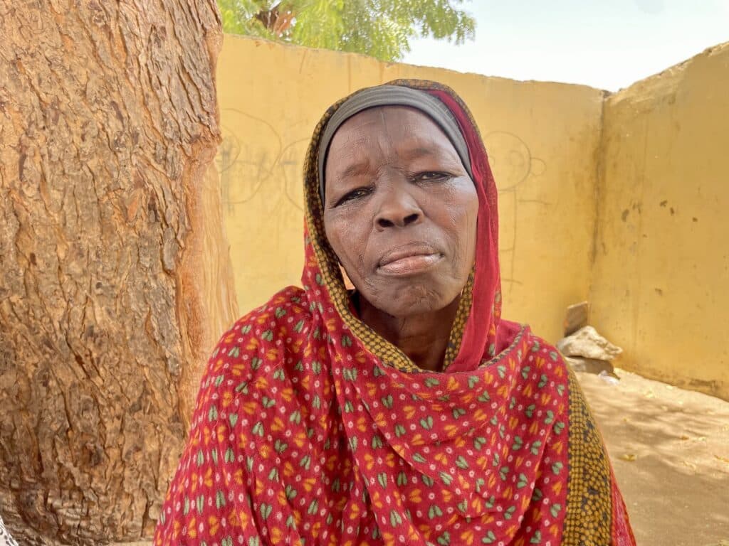 Elderly woman in a red patterned shawl leaning against a tree with a yellow wall in the background.