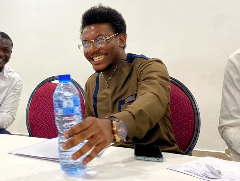 A smiling man with glasses reaching for a water bottle at a meeting, another person partially visible, with a phone and papers on the table.