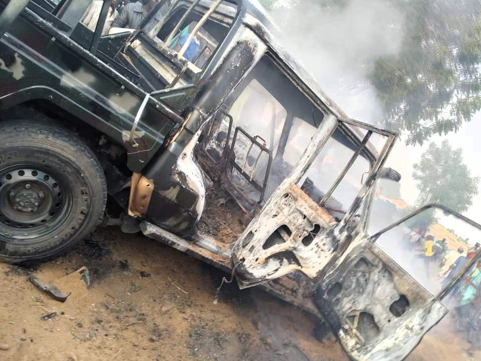 A burnt vehicle with open doors, smoke rising, and onlookers in the background.