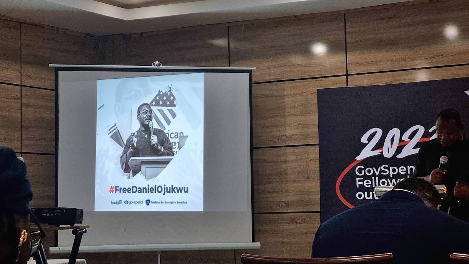 A projector screen with a man's image and text #FreeDanielOjukwu at a conference venue with people seated.