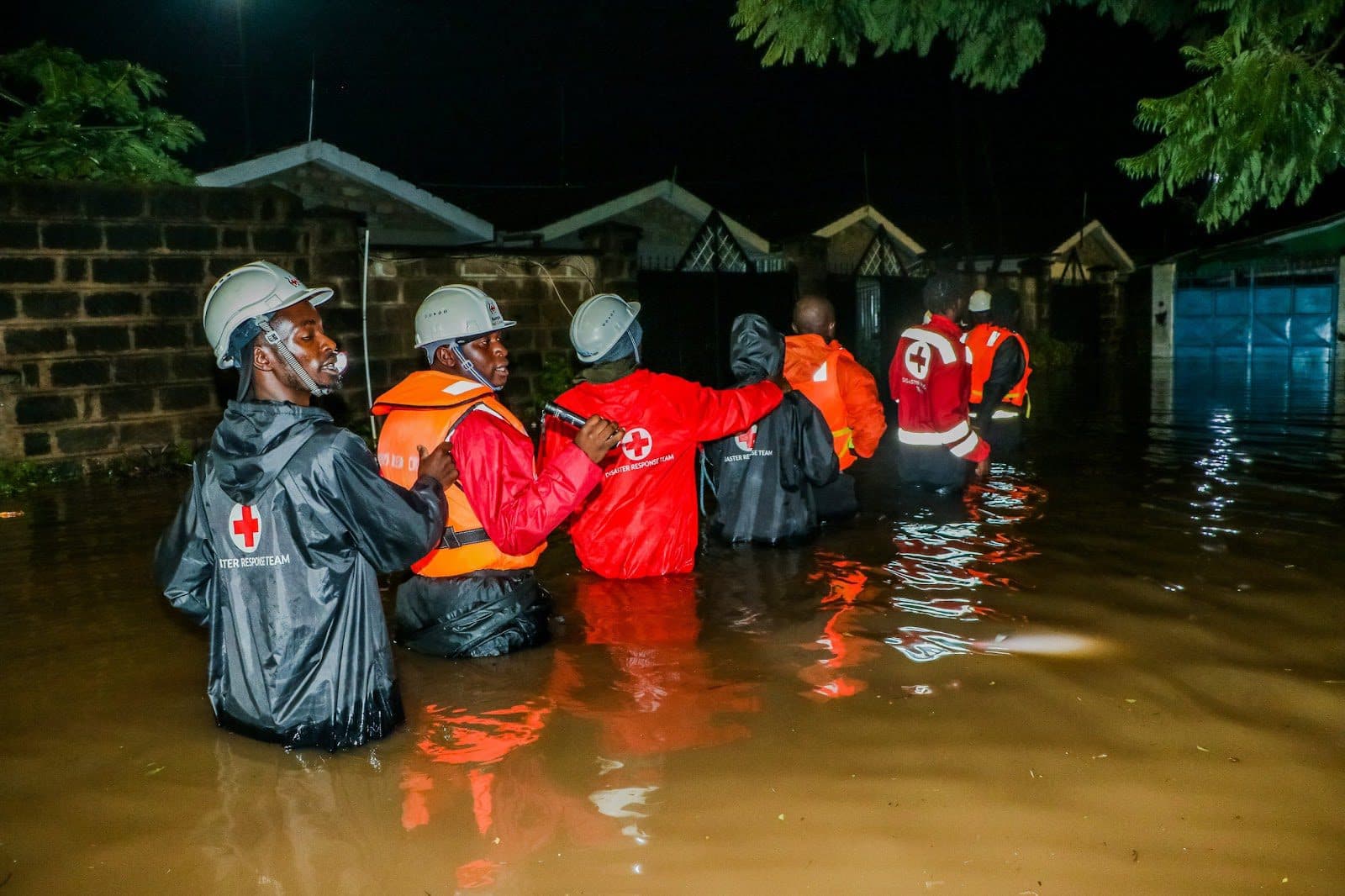 Rescue workers in reflective vests wading through floodwaters at night.