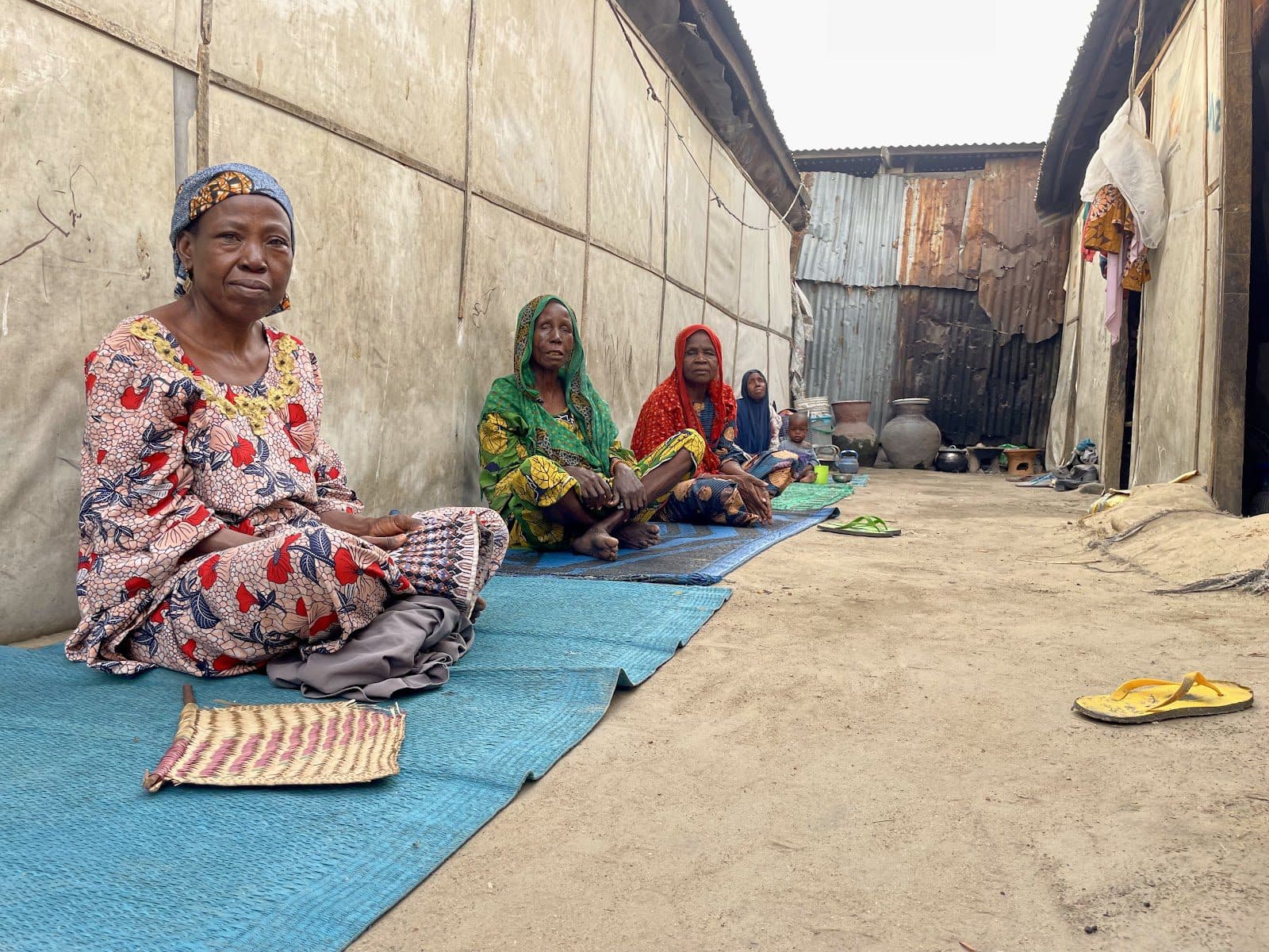 Women in traditional attire sitting outside on mats in a village alleyway.