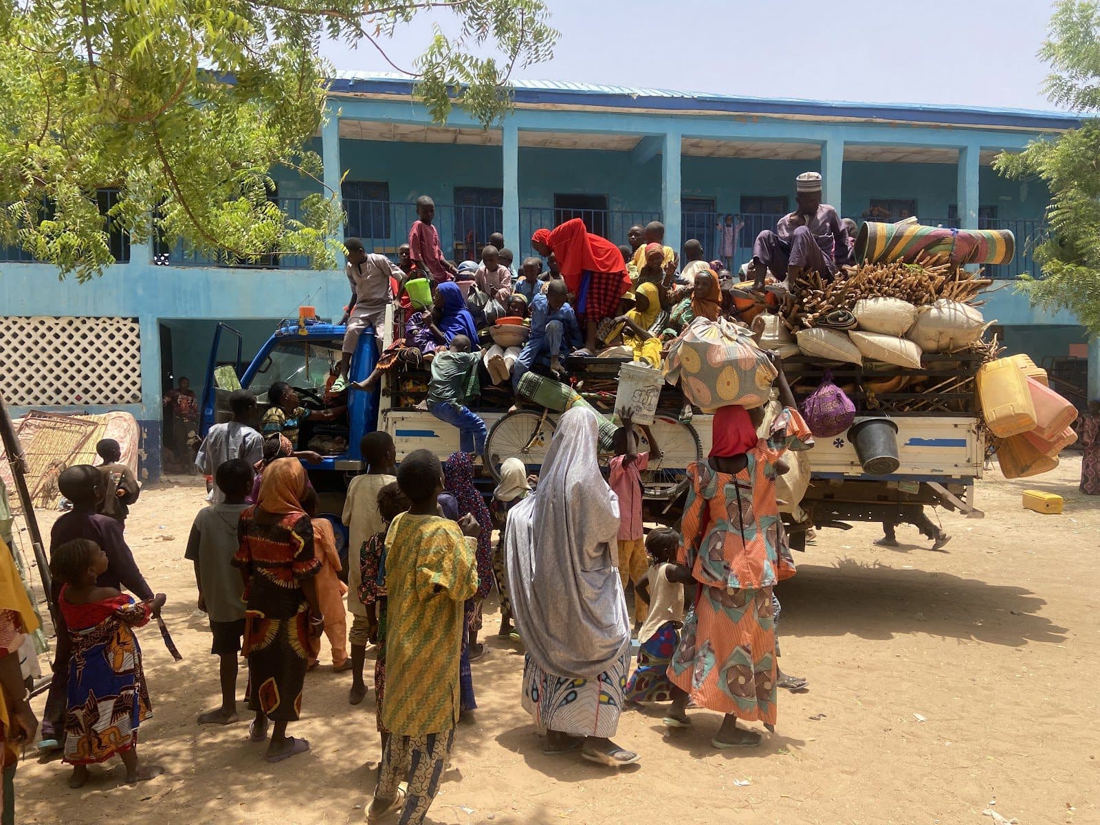 People and goods crammed atop and around a truck in front of a blue building.