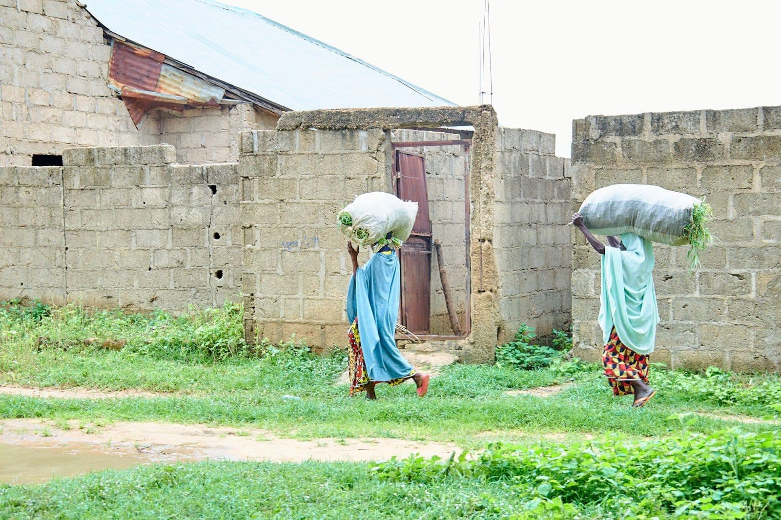 Two people carrying large sacks on their heads, walking by unfinished buildings.