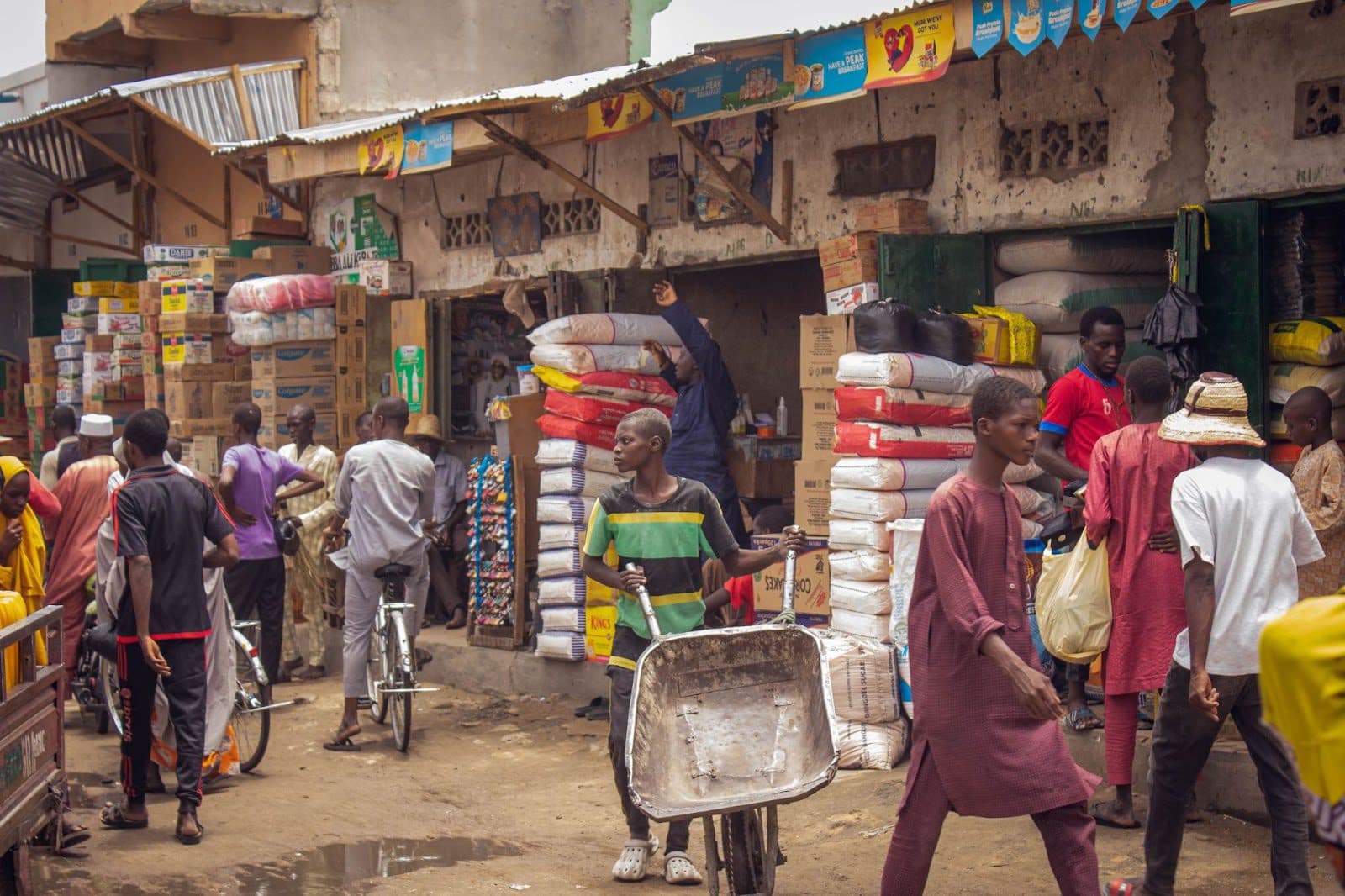 Bustling market scene with people shopping and transporting goods near stacked merchandise.