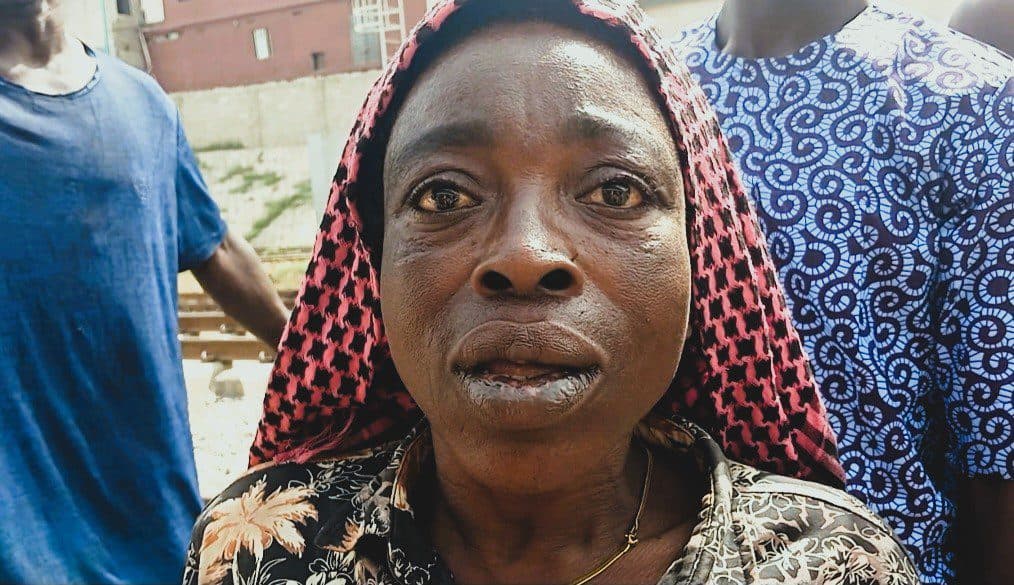 Elderly woman with a red headscarf, looking at the camera with people in the background.
