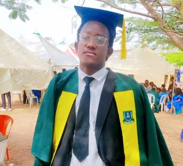 Graduate in cap and gown standing outdoors with celebratory tents and chairs in the background.