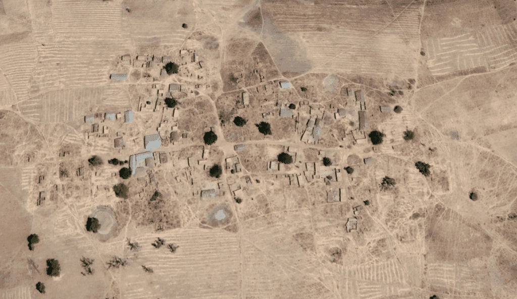 Aerial view of a sparse village with small buildings and trees amid dry, patterned fields.