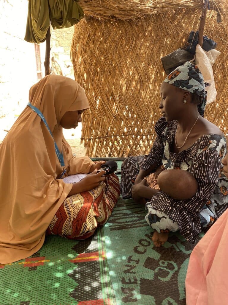 Two women in traditional clothing sit facing each other inside a hut, one holding a baby and the other with a notebook.