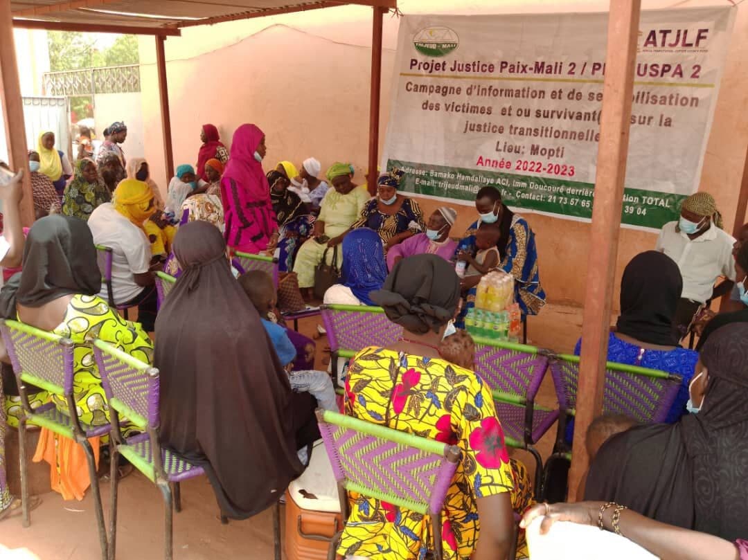 A group of people in colorful attire seated at an outdoor community meeting under a shelter.