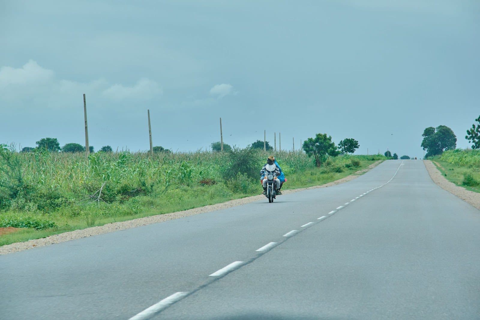 Motorcyclist riding on a rural road with greenery on both sides under a cloudy sky.