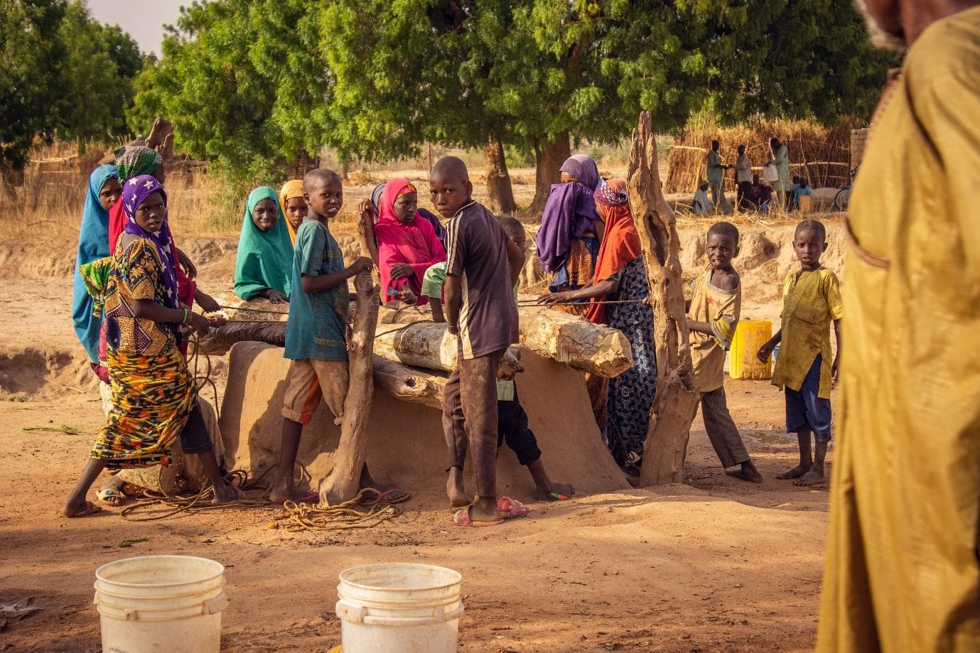 Group of people around a well with buckets, in a rural setting under a clear sky.