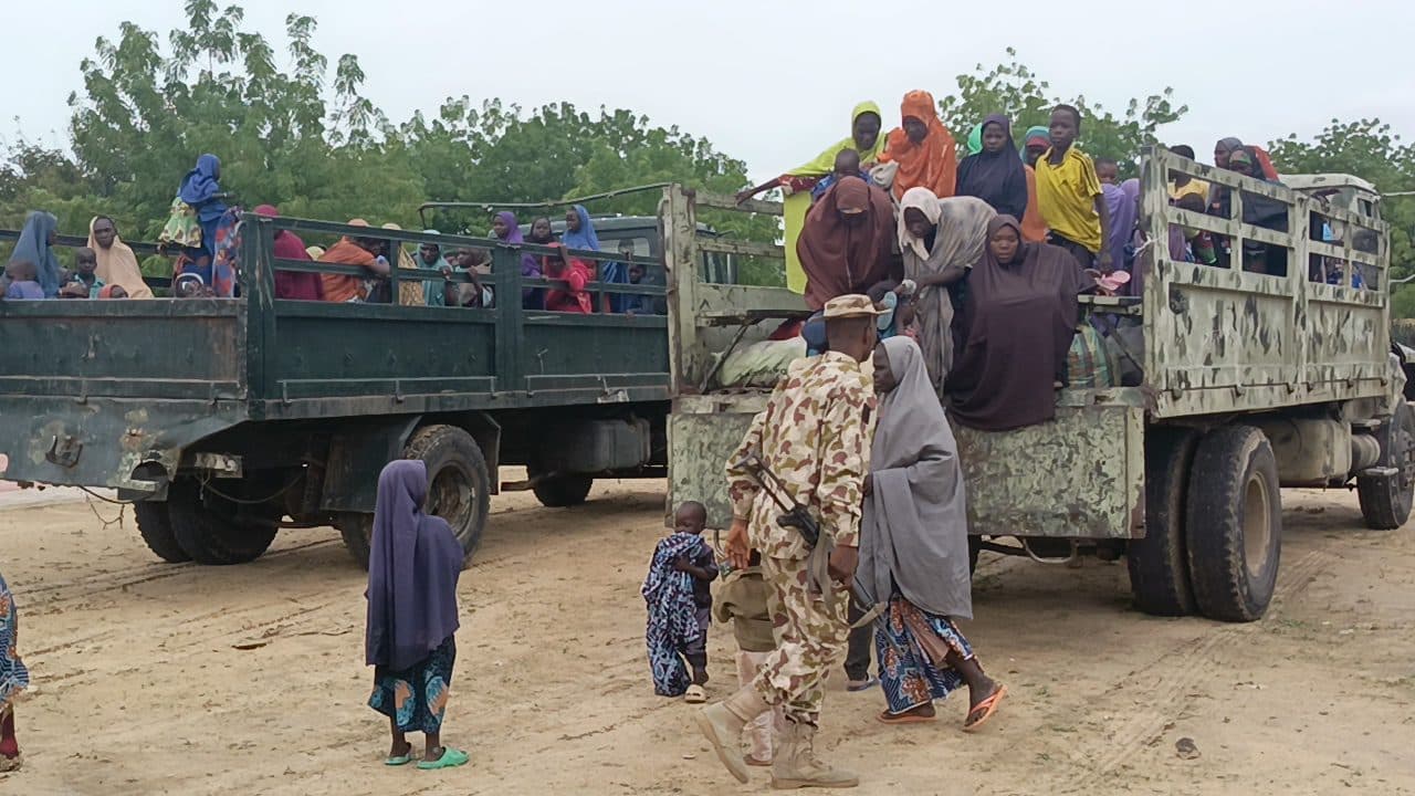 People in colorful clothing riding in open-back trucks with a soldier in the foreground.