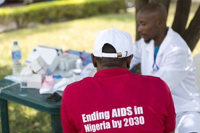 Man in a red shirt with "Ending AIDS in Nigeria by 2030" text, facing a healthcare worker at a table with medical supplies.