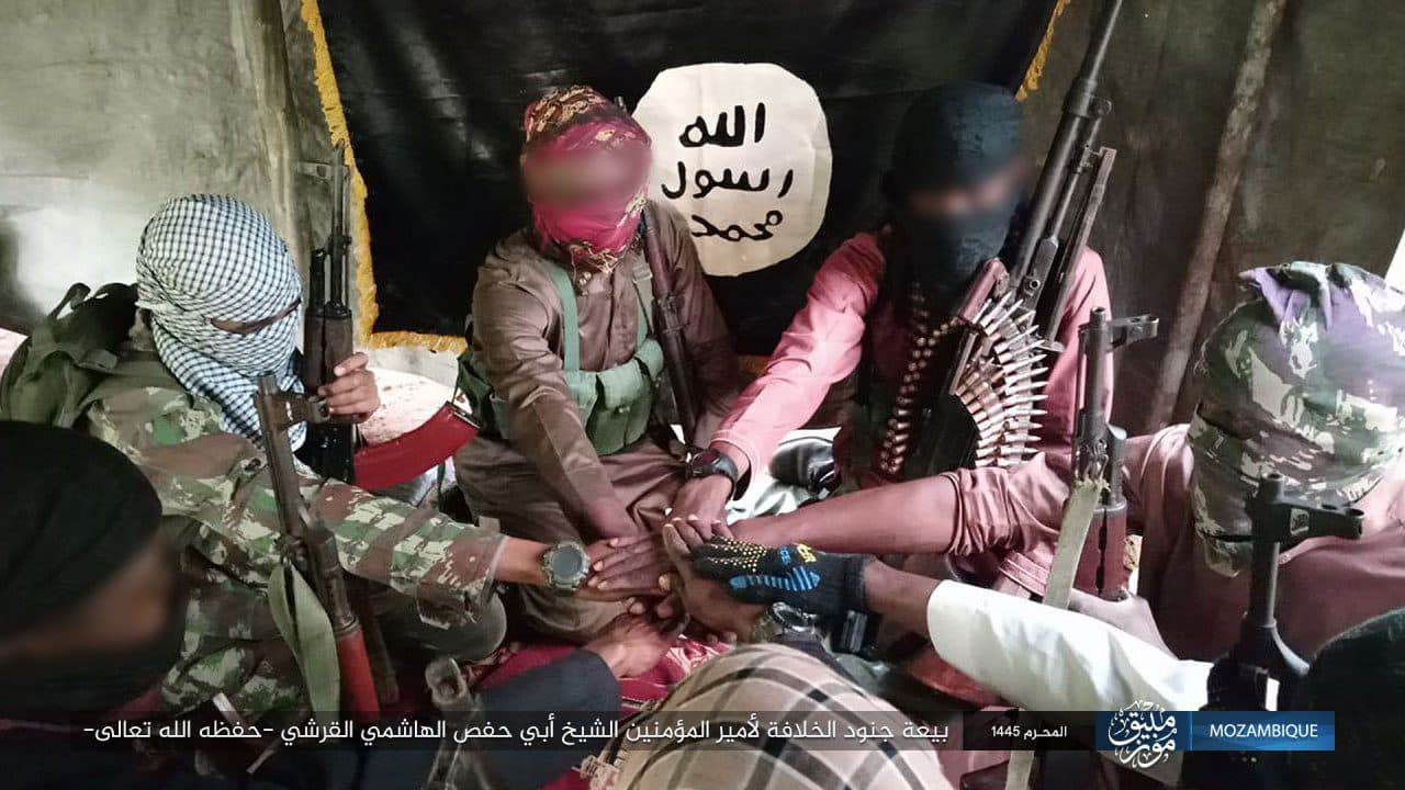 Group of masked individuals with weapons sitting together, with a flag in the background. Faces blurred for privacy.