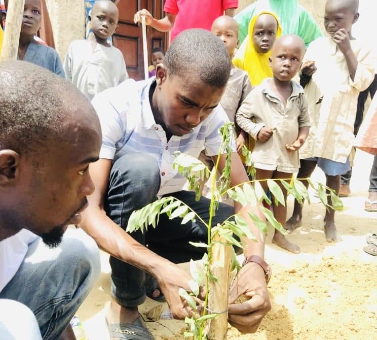Two men planting a tree with curious children watching in the background.