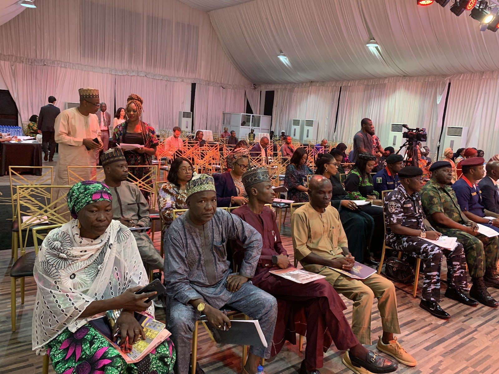 Attendees seated in a conference hall, some in traditional attire, with a videographer recording the event.
