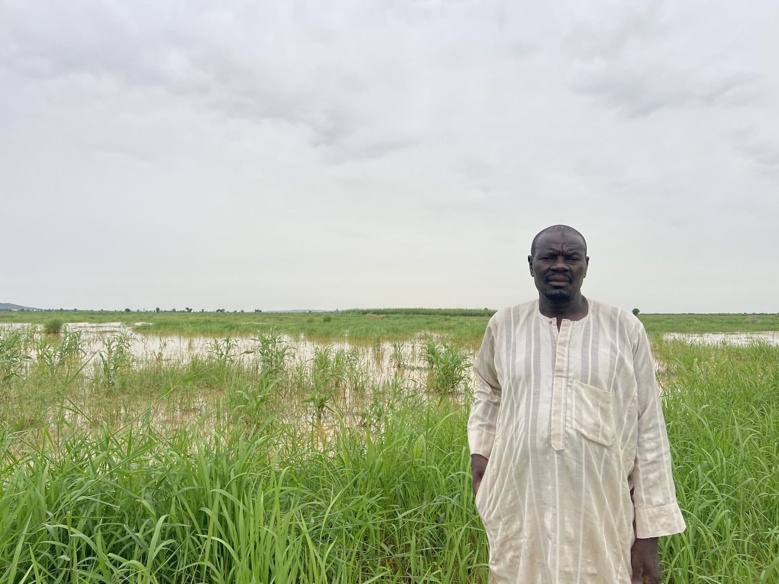 Man in traditional attire standing before a green field with a cloudy sky.