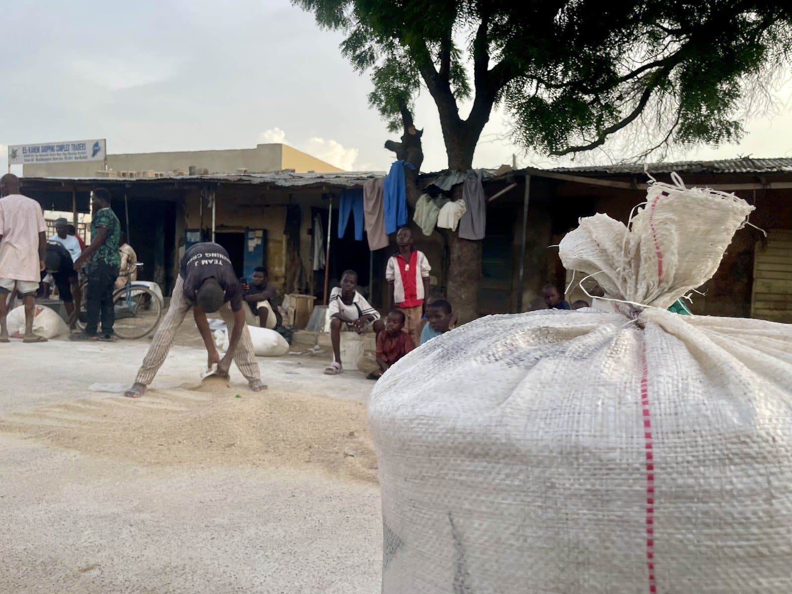 People in a market with large bags in the foreground and a tree providing shade.