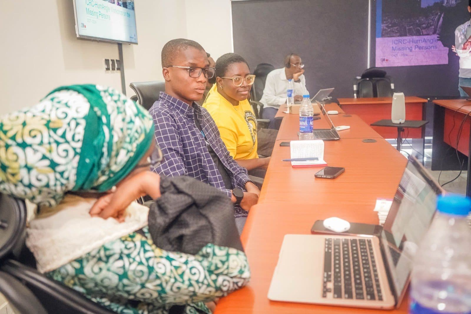 Three individuals focused on a presentation in a conference room with laptops and personal items on the table.
