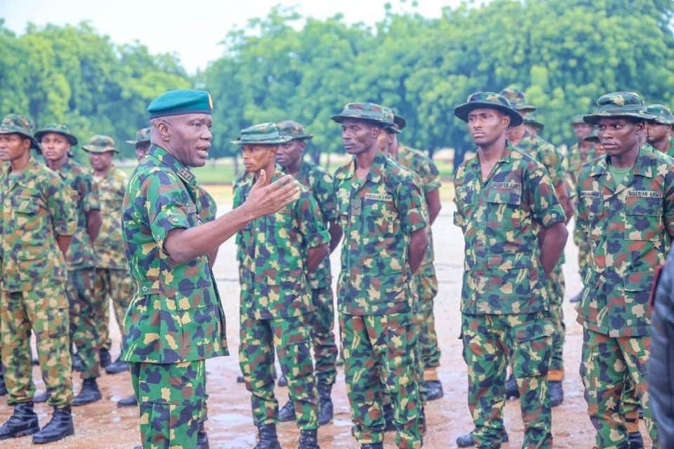 A group of soldiers in camouflage listening to an officer speaking outdoors.