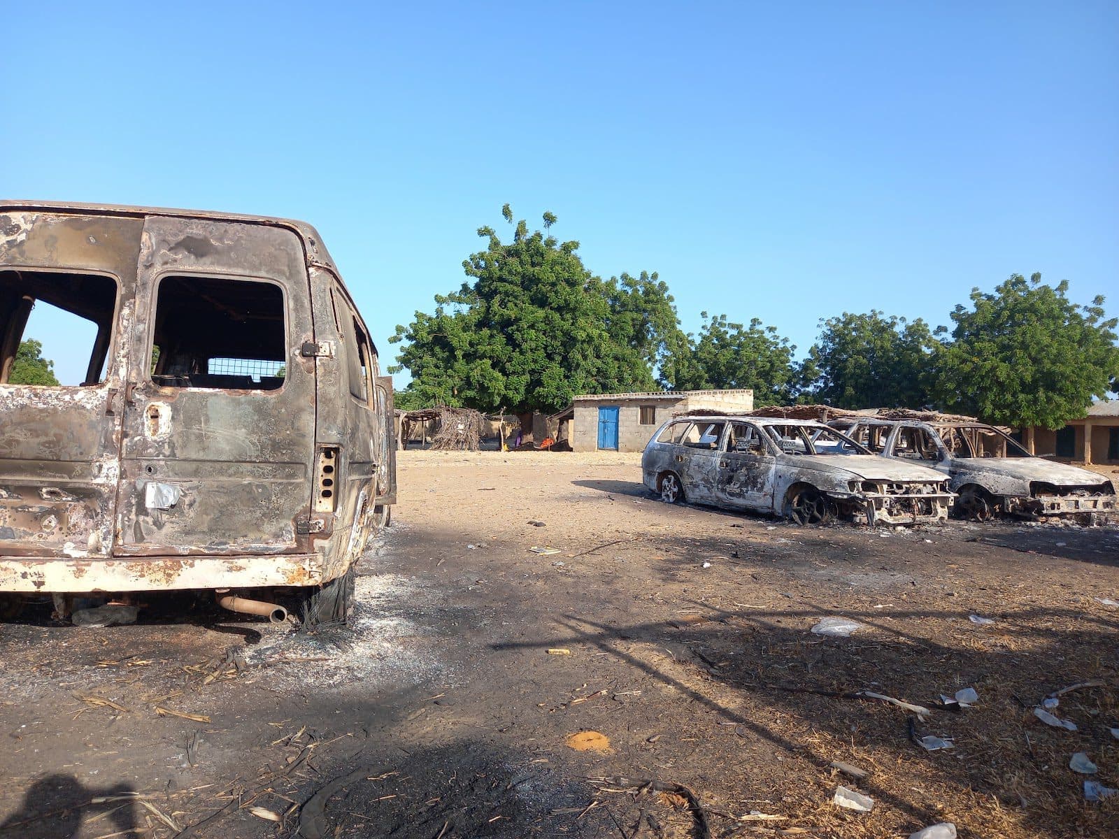 Burnt-out vehicles in a dusty lot with trees and buildings in the background.