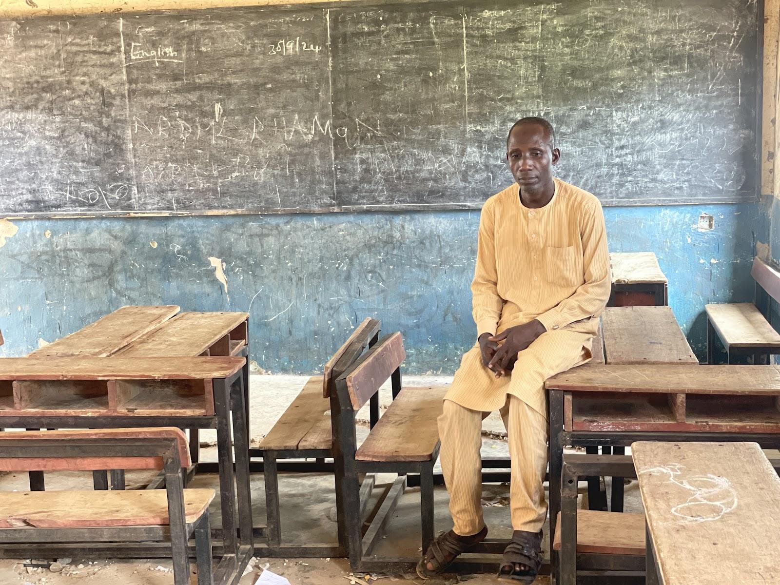 Man seated in a classroom with wooden desks and a chalkboard behind him.