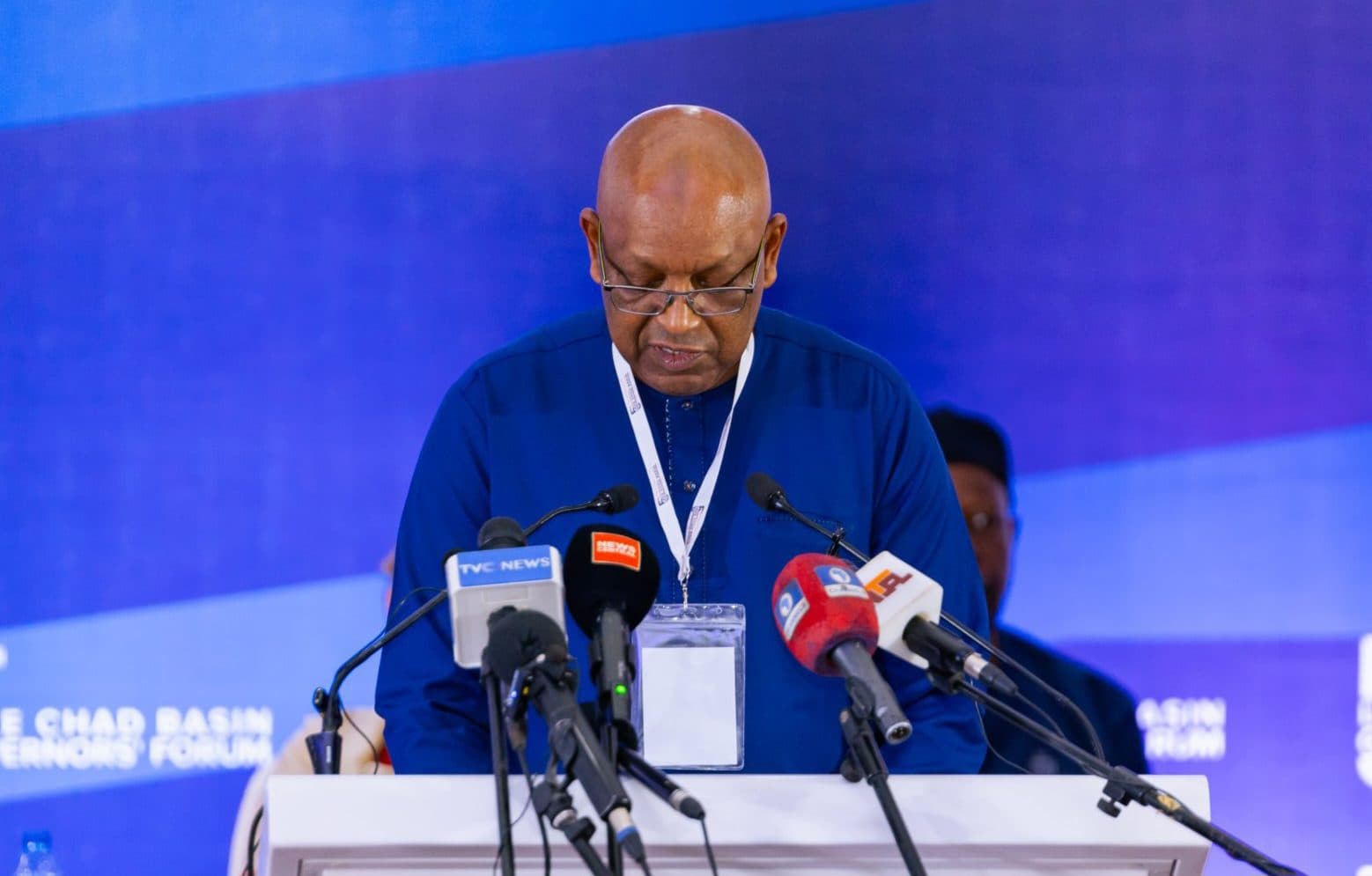 A person in a blue shirt speaks at a podium with multiple microphones against a blue backdrop at a conference.