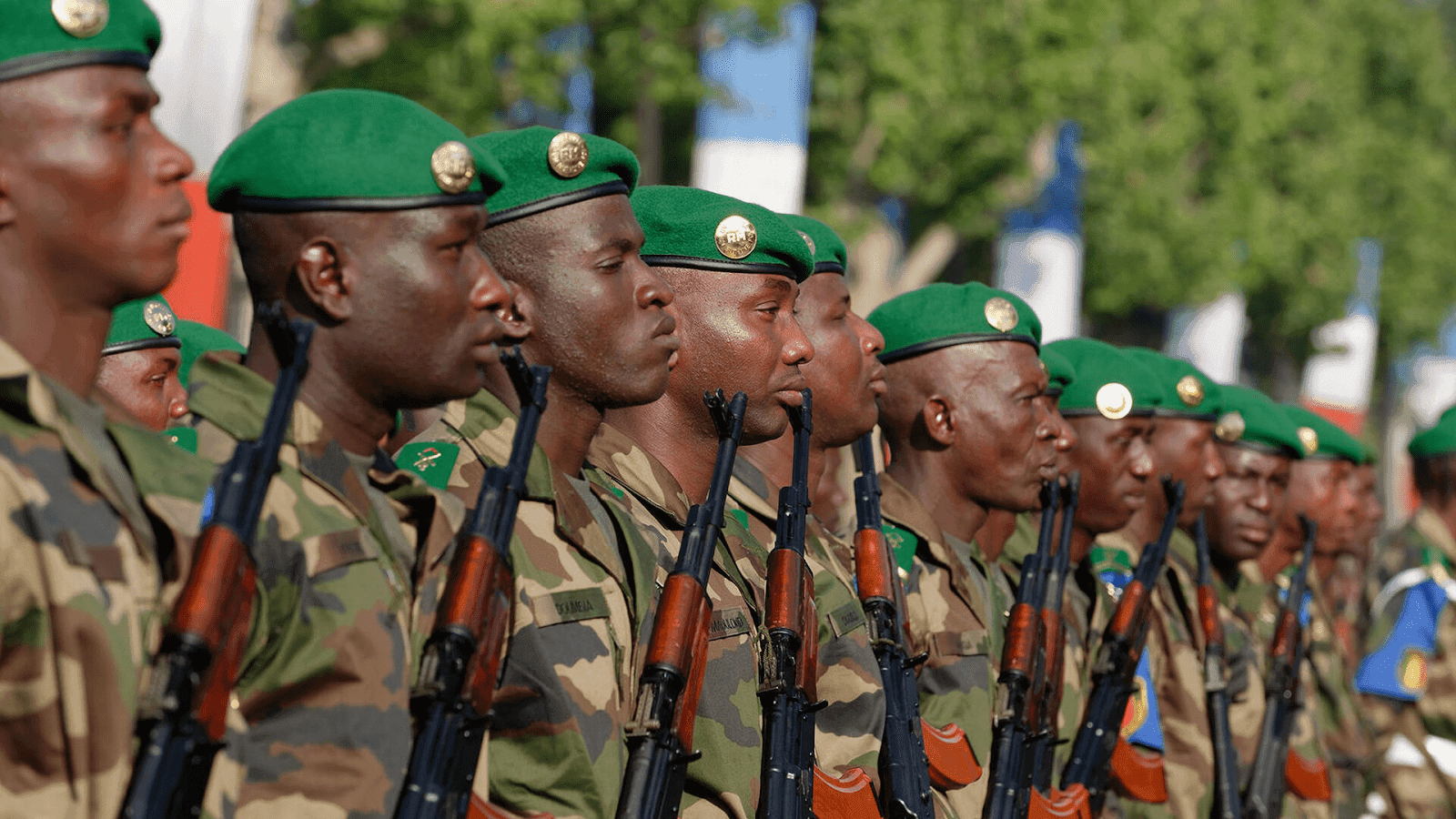 Soldiers in green berets and camo uniforms stand in formation, holding rifles, with trees in the background.