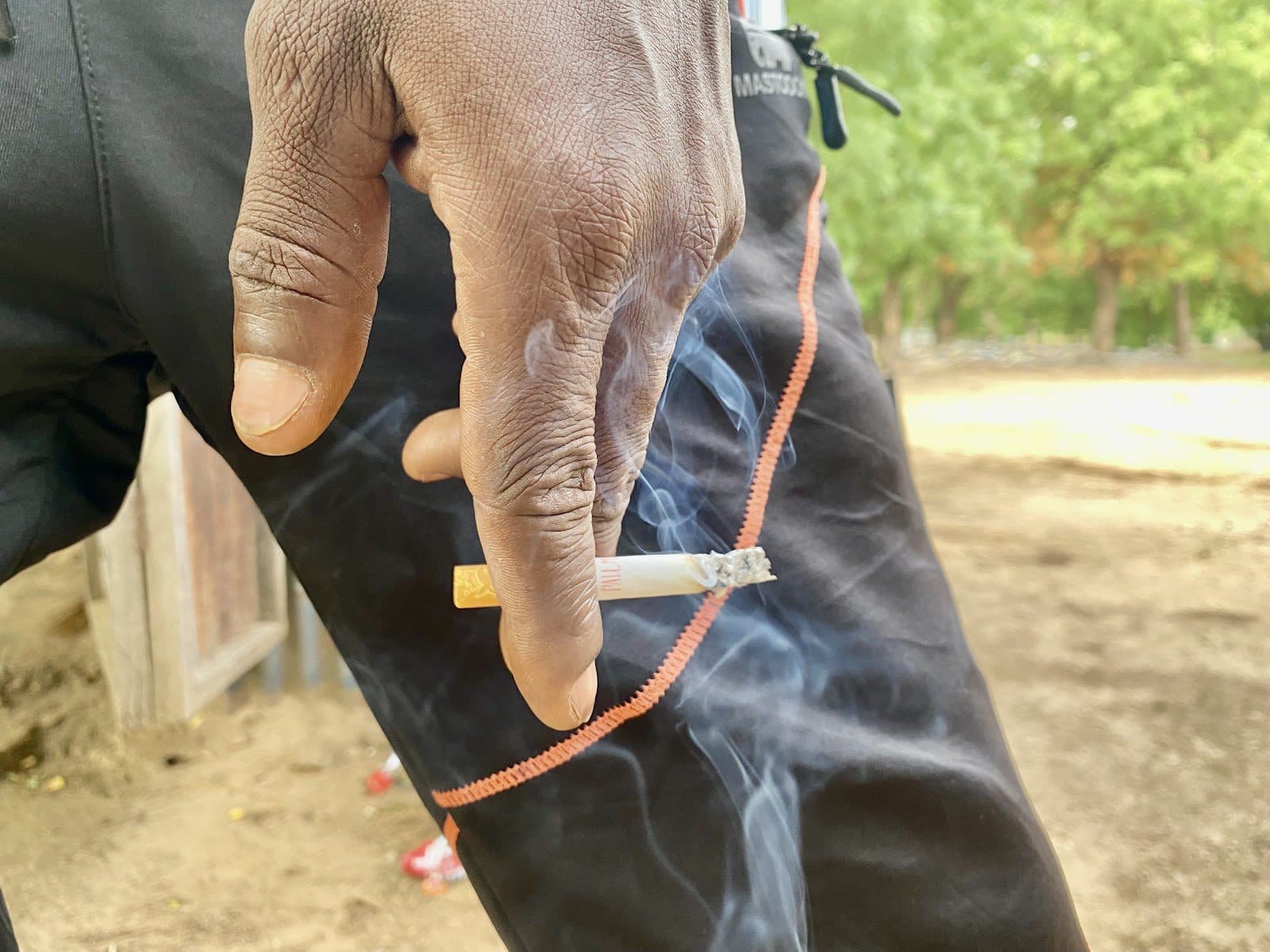 Hand holding a lit cigarette against black pants with orange trim, outdoors. Smoke wafts in the air.