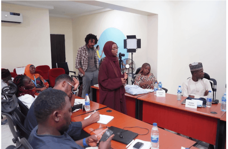 A person in a maroon hijab is speaking at a conference table. Several attendees are seated, listening and taking notes.