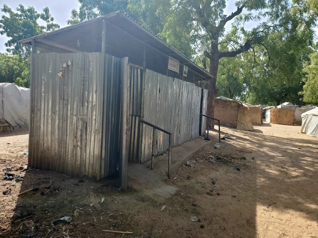 Corrugated metal structure next to trees under clear sky, with several huts and tents nearby on sandy ground.